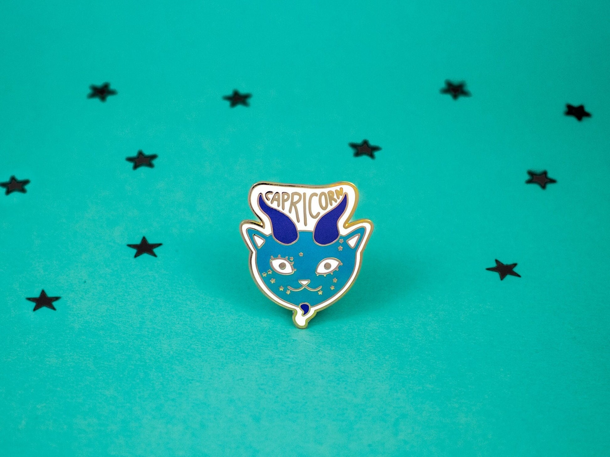 Capricorn cat enamel pin in turquoise colour with horns and a goatee and capricorn constellation speckled across its face