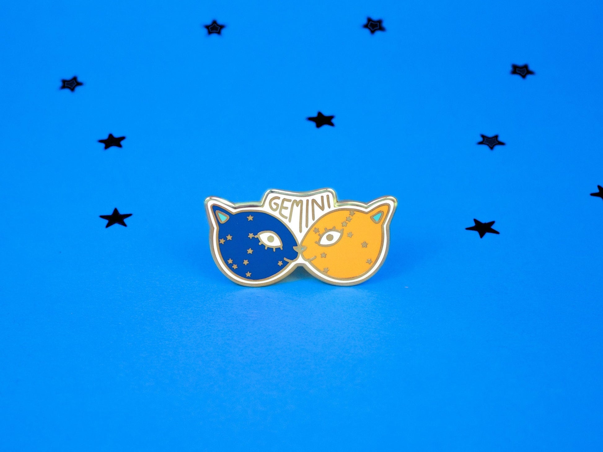 enamel pin of two kitties touching noses, one dark blue and one pale orange with the word gemini and the gemini constellation speckled across their faces