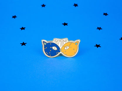 enamel pin of two kitties touching noses, one dark blue and one pale orange with the word gemini and the gemini constellation speckled across their faces