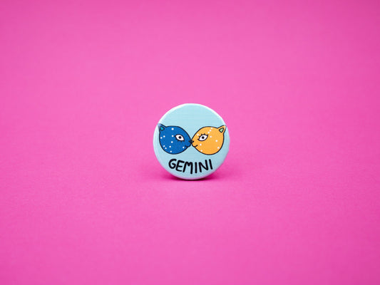 blue button badge with two cats, one blue and one orange, face to face with the gemini constellation across their faces and the word &#39;GEMINI&#39; below